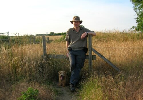 An older man wearing a hat stands in front of a style. At his feet is a Norfolk Terrier. Behind him are golden autumn fields.