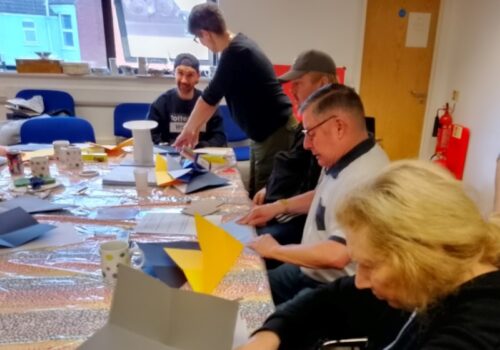 participants of a creative workshop are sitting at a long table. They are intently folding paper to create origami shapes