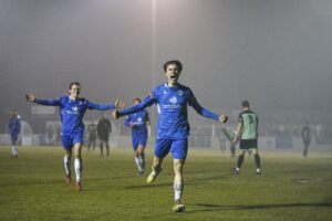 Jubilant football players, dressed in blue kit, run towards the camera whilst the background is shrouded in mist