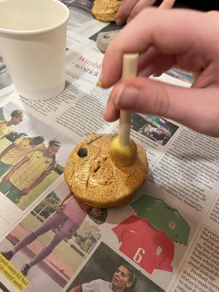 Close up of a hand holding a small stick with sponge, applying gold paint to a small disc with a hole pierced in it.