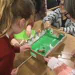 A cardboard football game is on the table. An adult and a child each has a stick running under the box, used to propel the magnetic football figure across the board. Another adult steadies the game