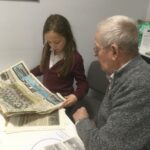 A gentleman seated beside a school girl, both looking at an old newspaper