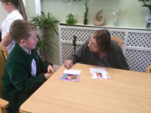 A young lad in a green jersey seated at a table, in conversation with a woman who is looking at a decorated card in front of her