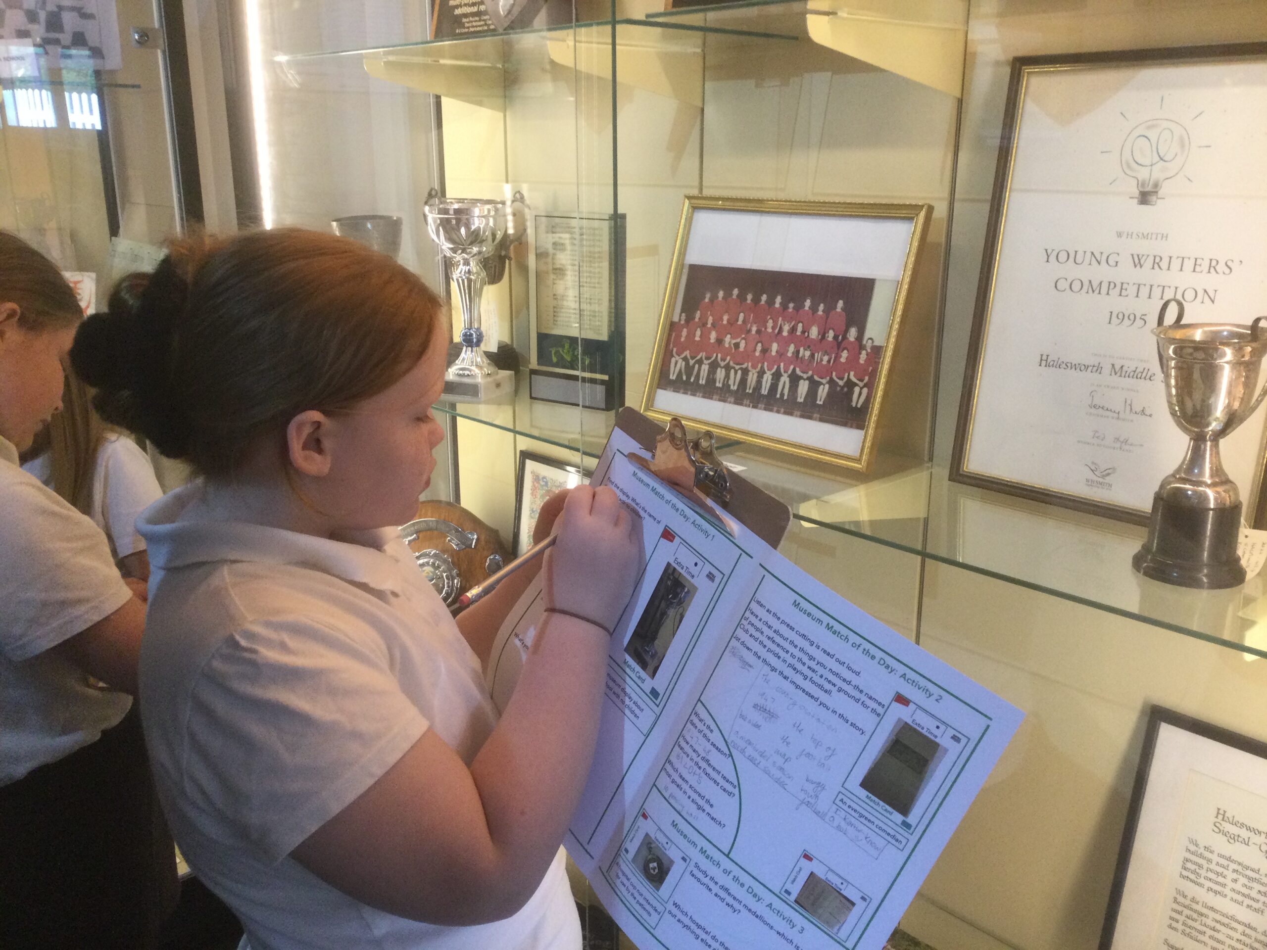 A school girl in front of a display case with trophies and photographs, writing something on her worksheet.