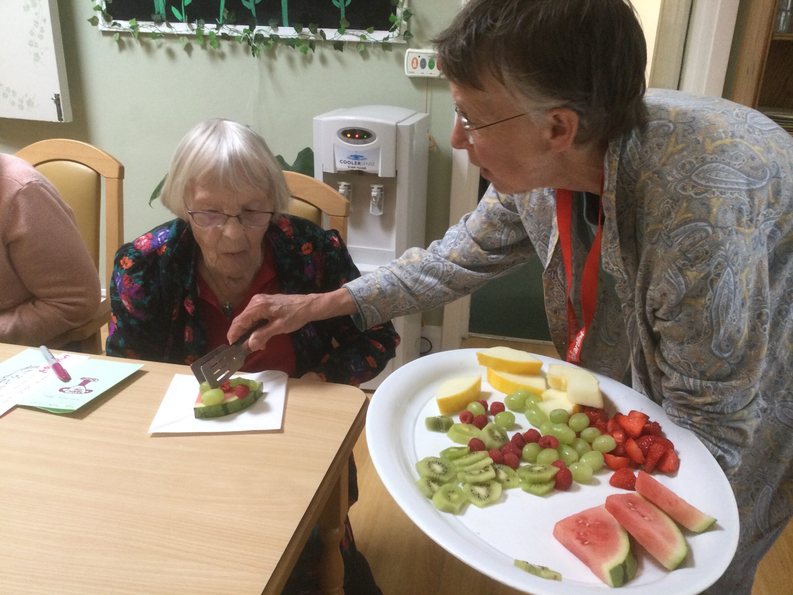A person with a large white platter with sliced watermelon, honeydew melon, grapes, kiwi, strawberries and raspberries, serving some fruit to a resident