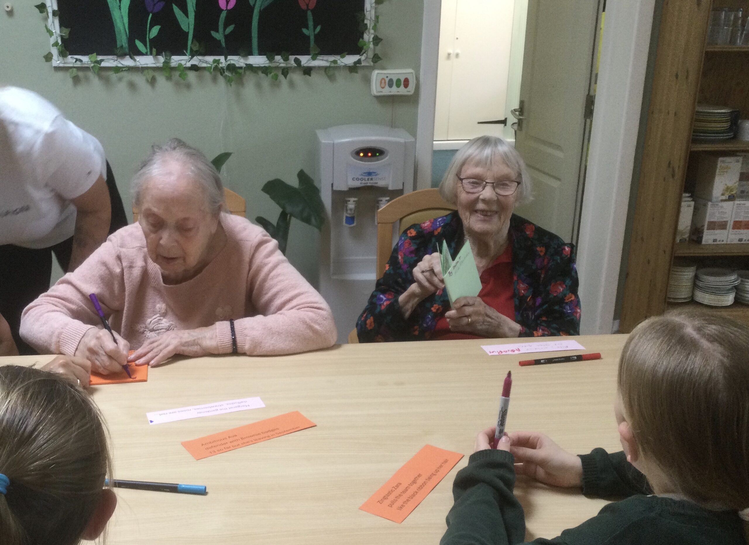 Two elderly ladies writing and reading handwritten booklets, seated opposite a pair of school children