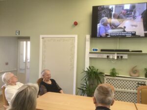 A group of people seated at a table watching a video on a large flat screen on the wall