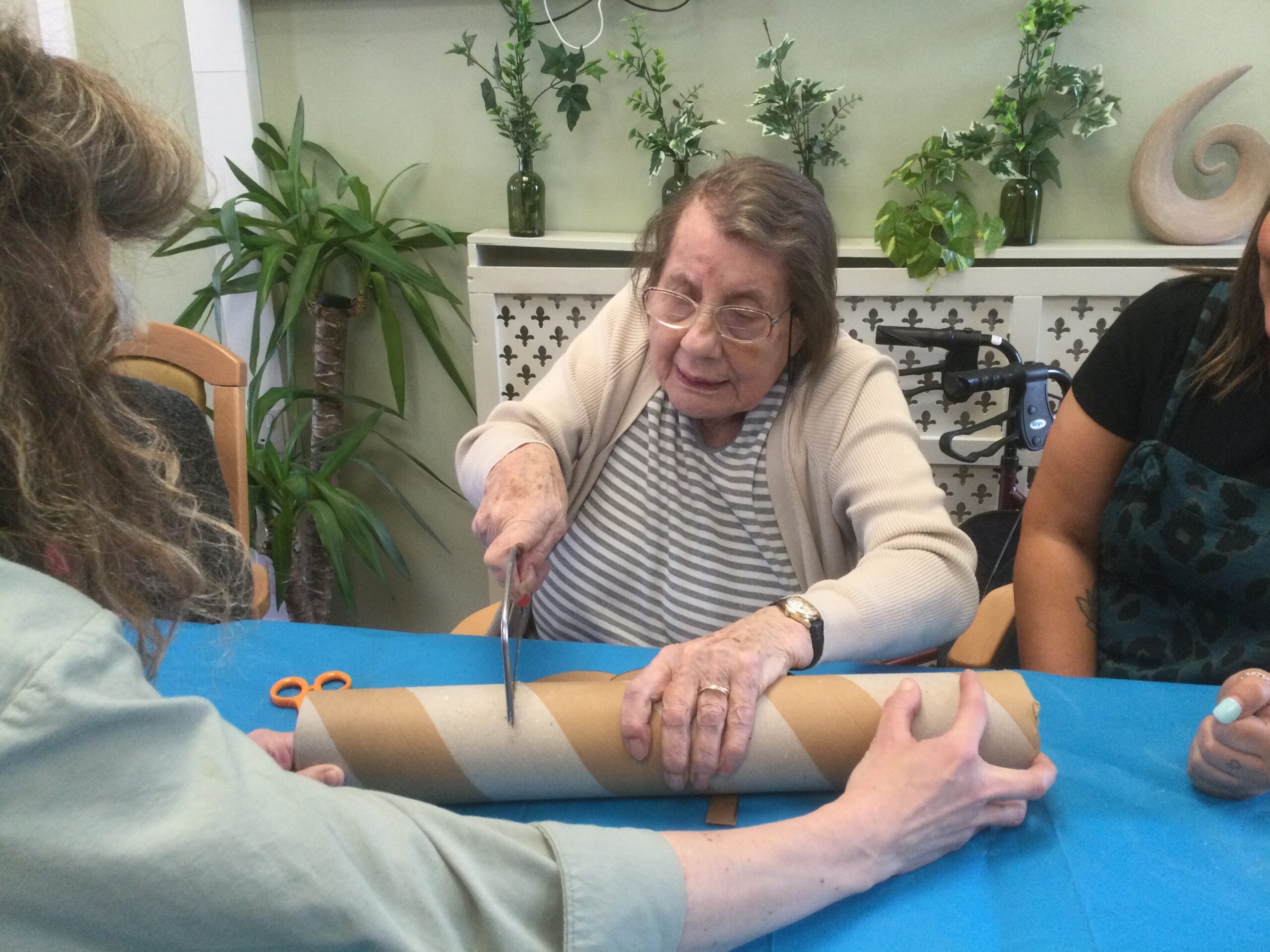 A woman seated at a table cutting through a cardboard tube whilst another person steadies it for her