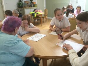 School girls seated at a table with two adults, with worksheets on the table in front of them - one child holds a magnifying glass