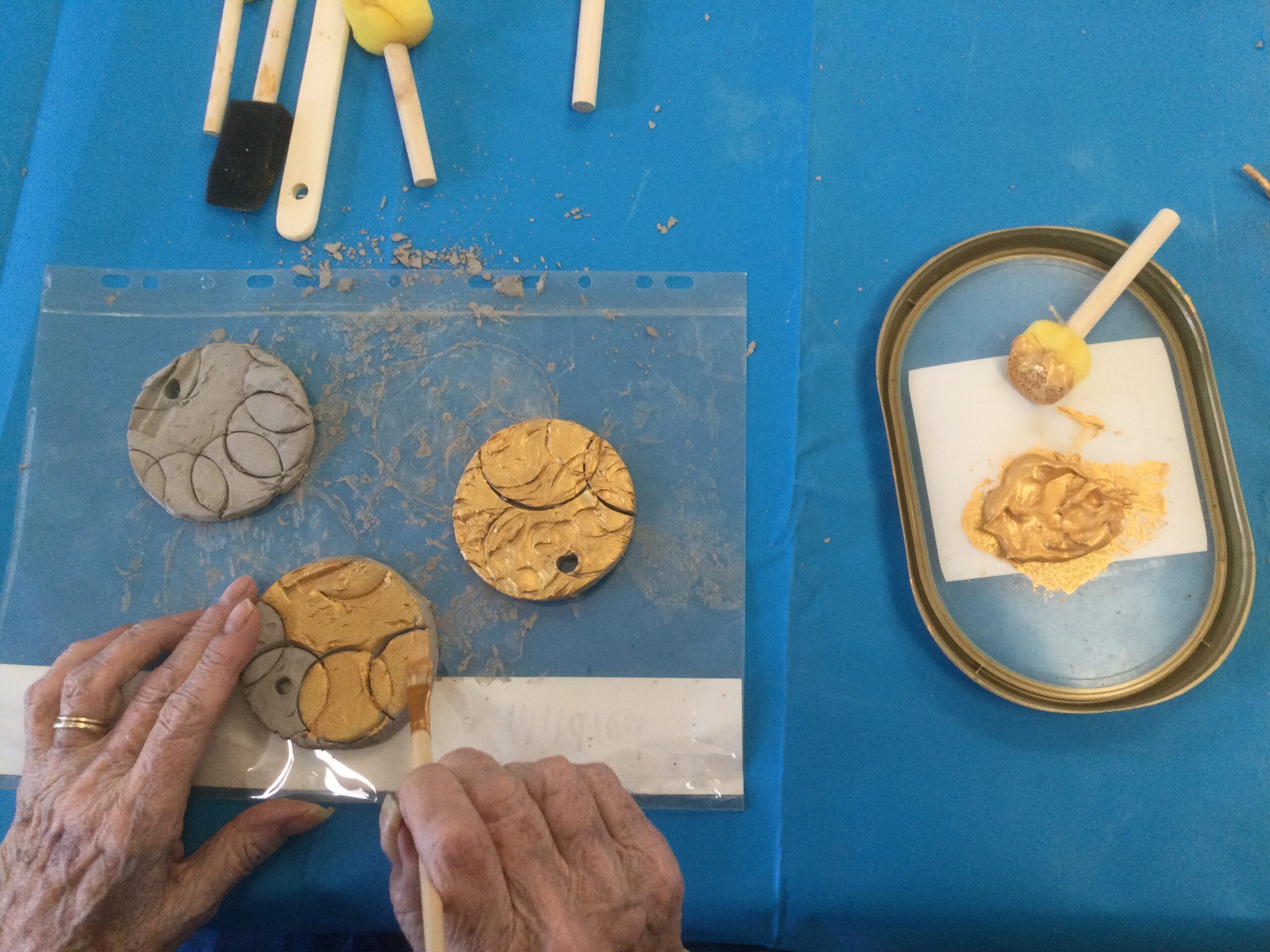 Overhead image of a pair of hands, some clay medals being painted gold. There's a plastic lid with gold paint and sponge applicator to the right and more applicators in front of the medals