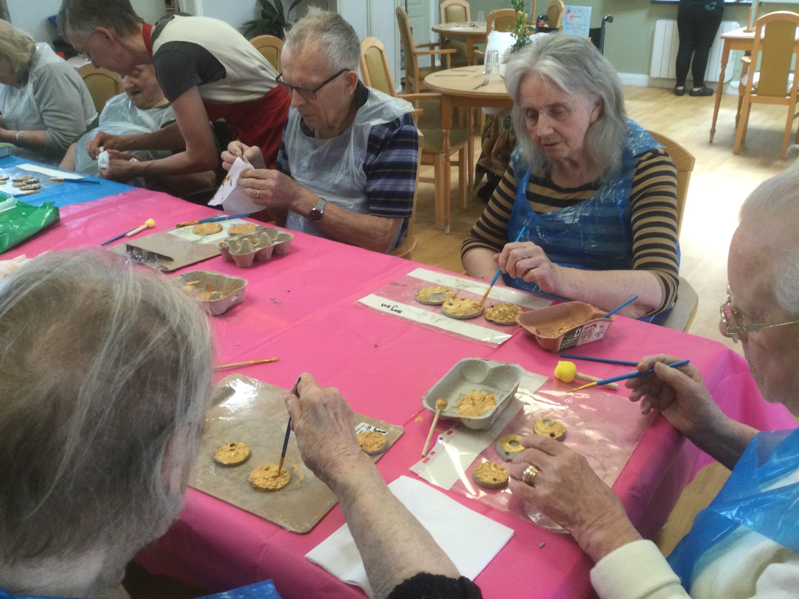 A group of people wearing blue plastic aprons seated at a table painting clay medals with gold paint