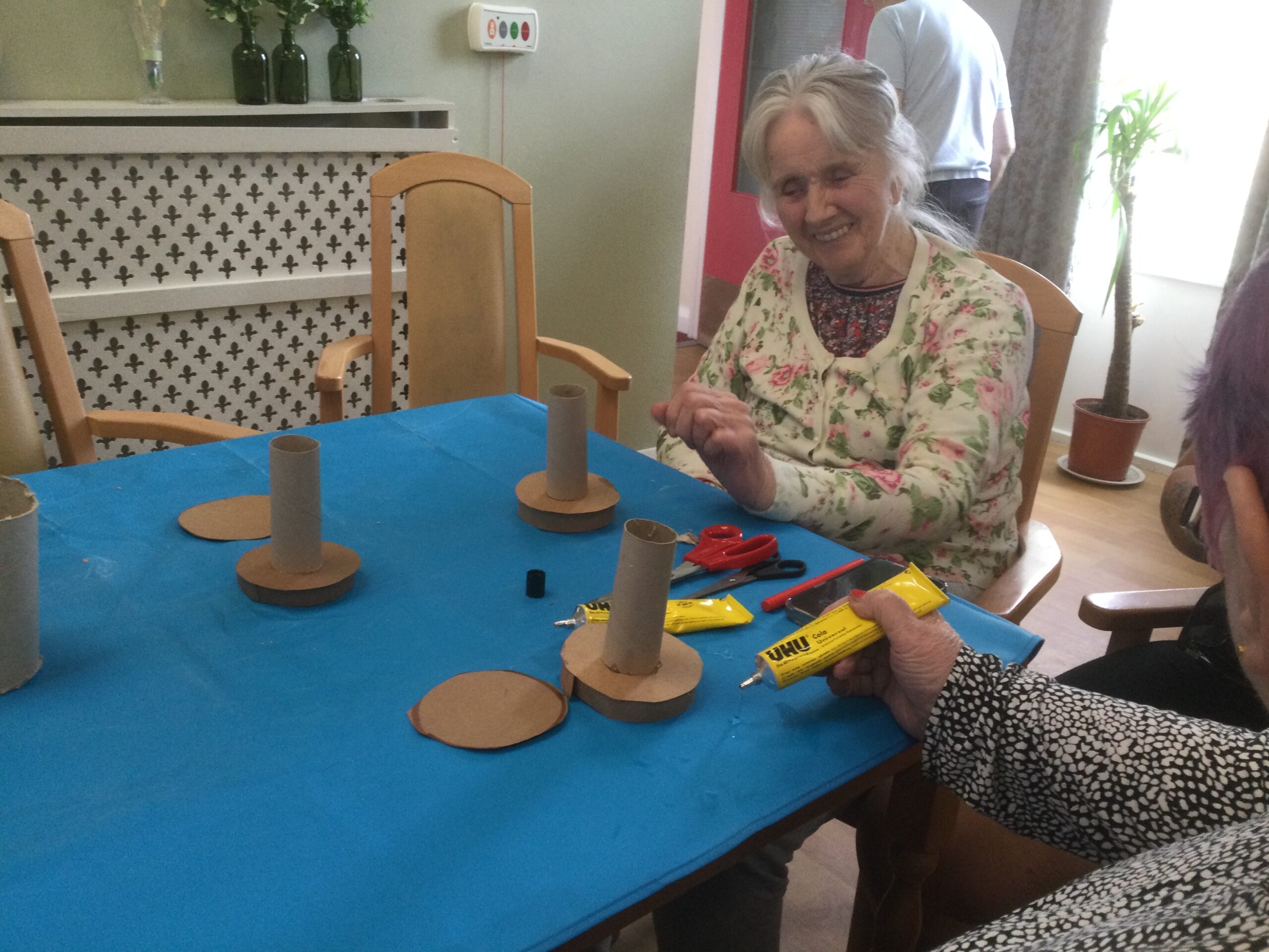 A woman smiles as she looks at an array of cardboard tubes and bases on the table in front of her. Another hand can be seen, holding a yellow tube of glue