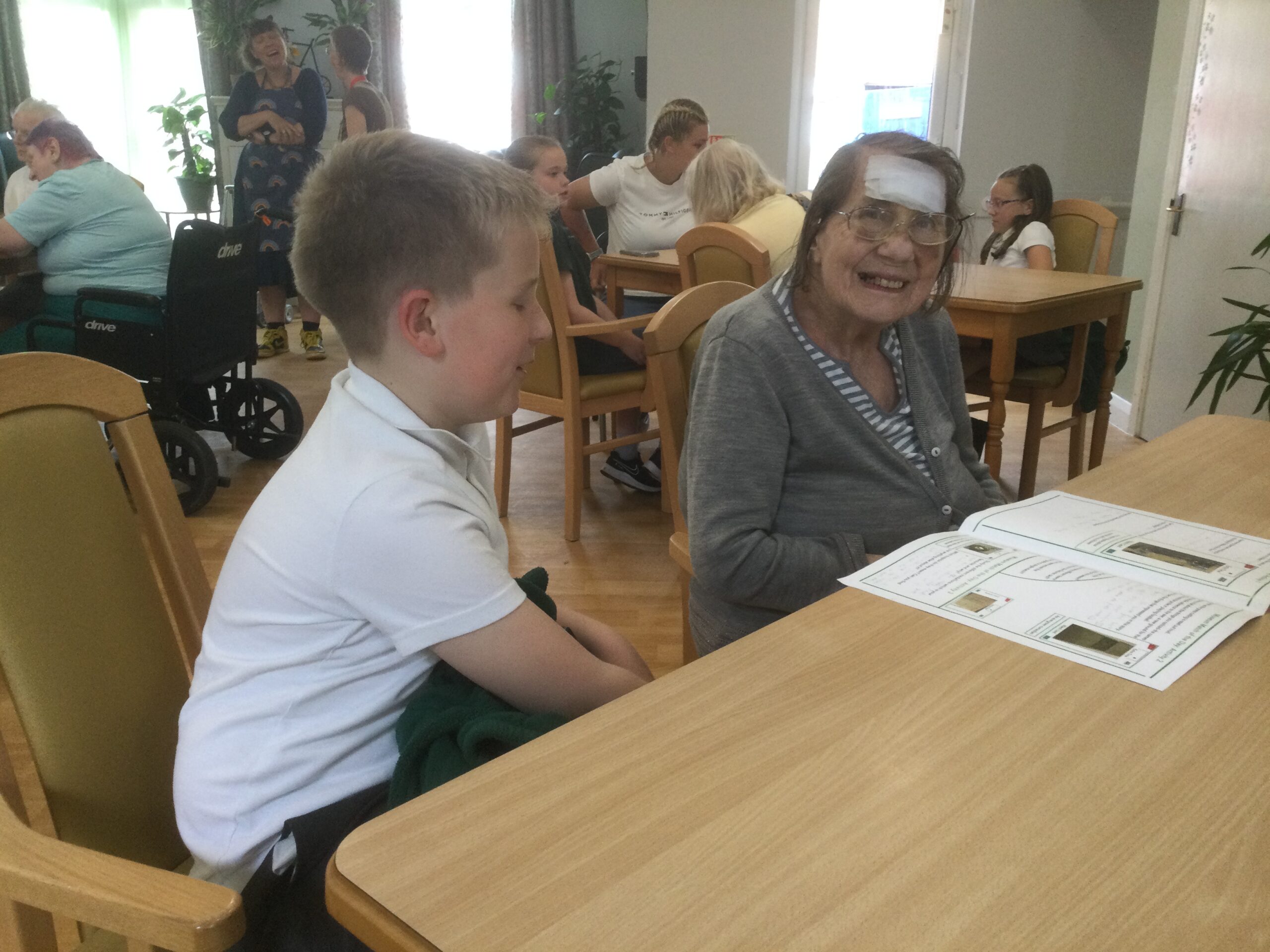 A woman with a large white bandage on her head seated beside a school boy discussing his work. Several more people in the background