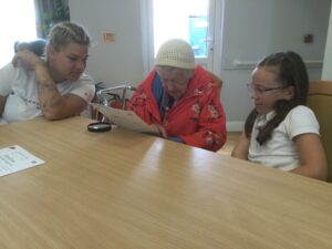 A woman in a red floral raincoat and woolly hat peers closely at a worksheet, with a child and adult seated either side of her