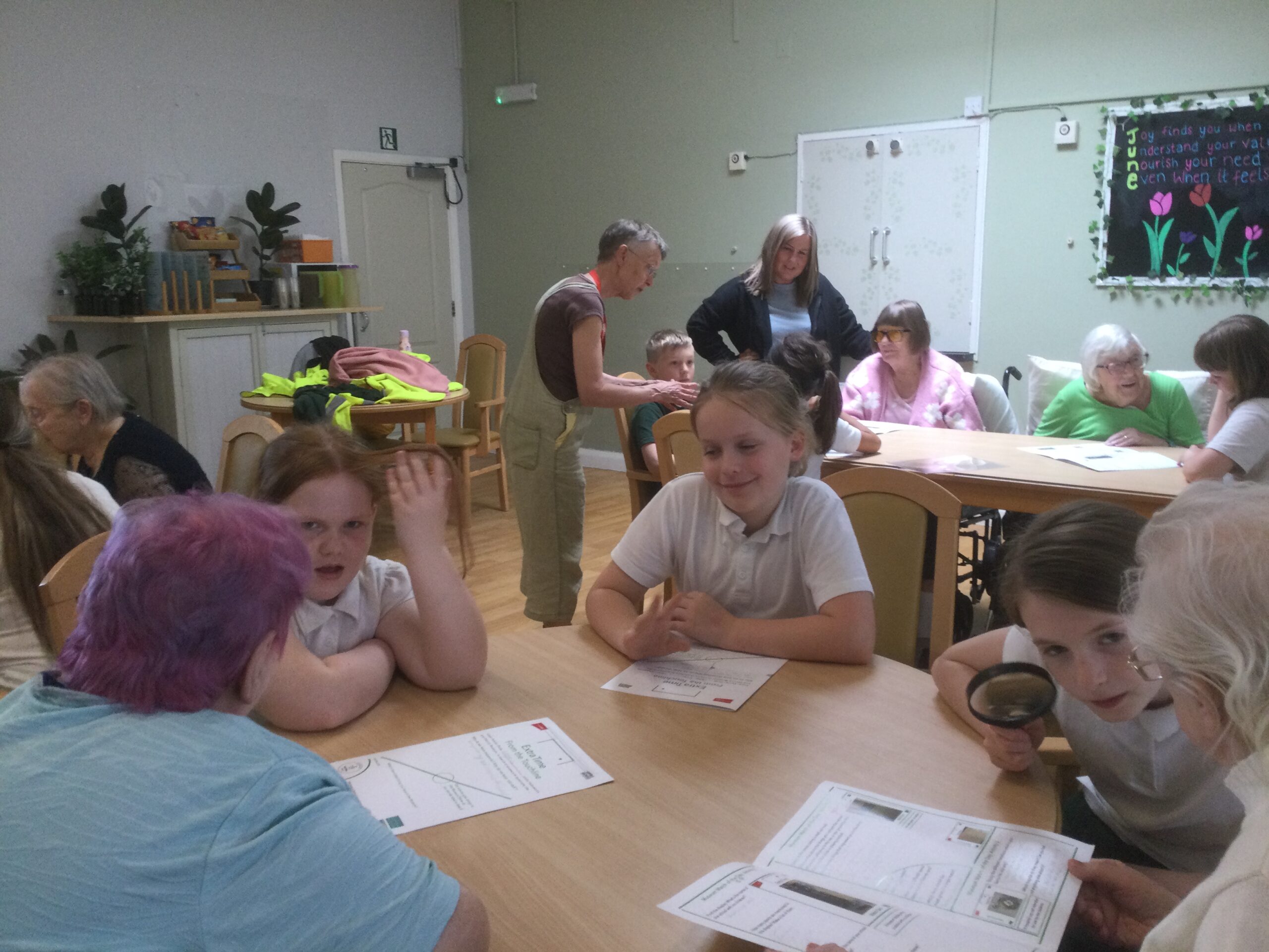 A room full of people, with children seated at table in the foreground and adults on another table in the background. There's a large blackboard on the wall with three pink flowers painted on it