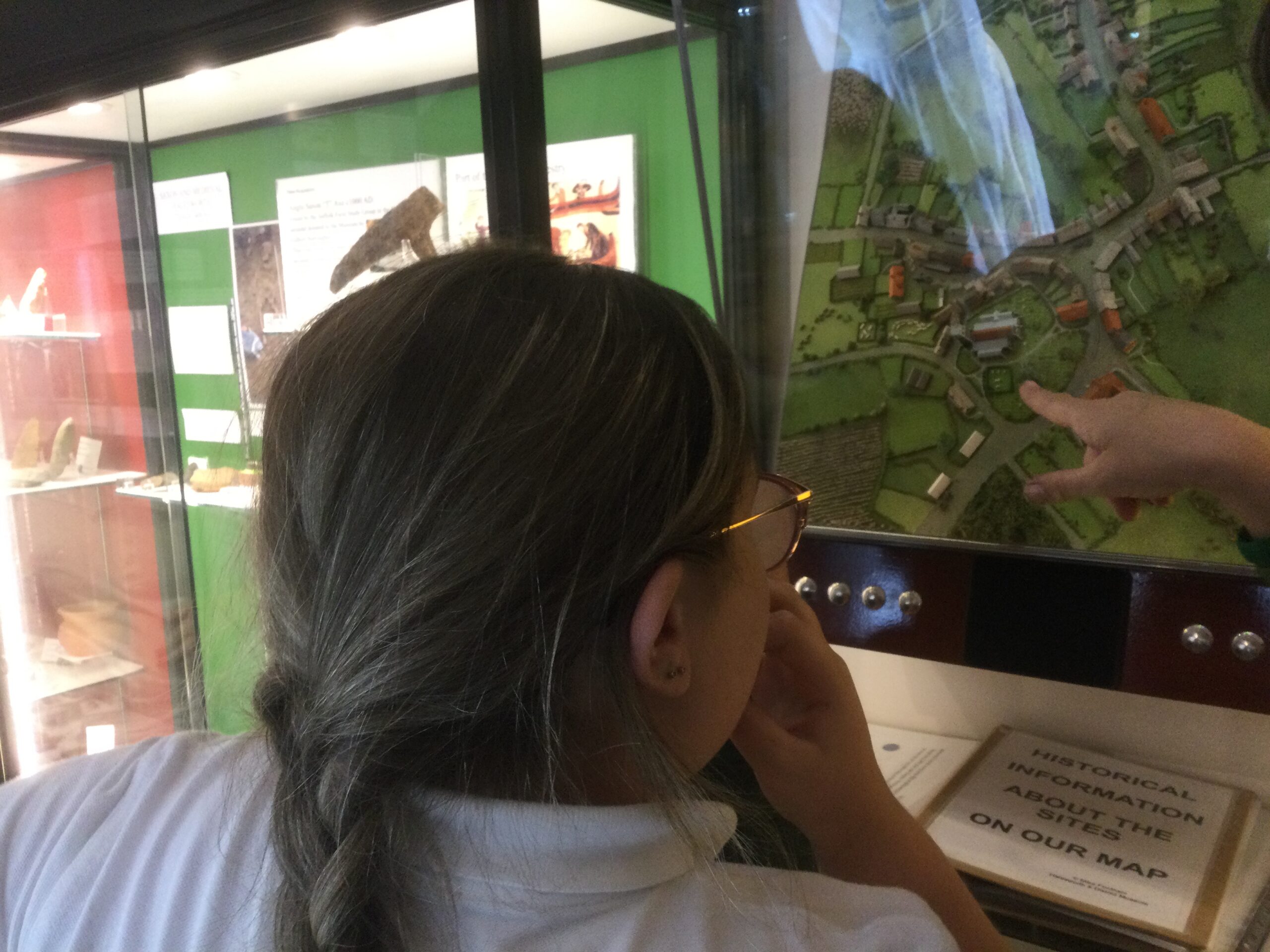 A child looks closely at a map, with a second child pointing out a particular spot. Light bounces off the glass case, making it difficult to see the map in detail.