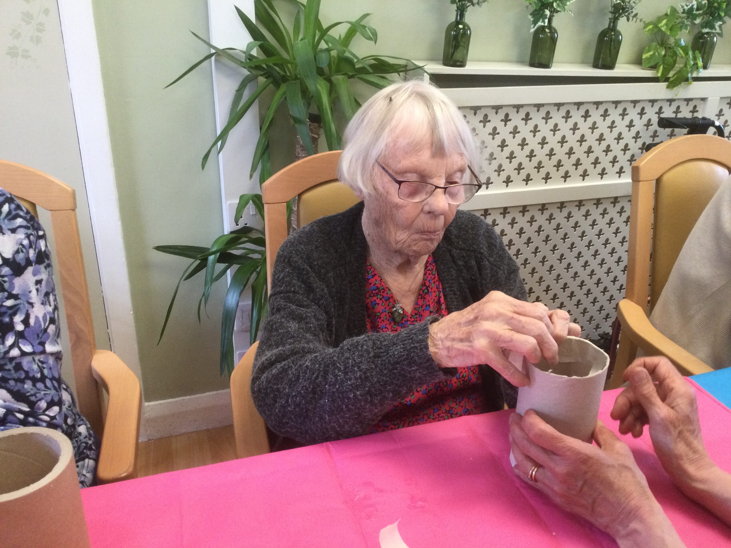 A woman seated at a table with plants on a shelf behind her. She is working on a piece of cardboard tubing that is being held by another pair of hands