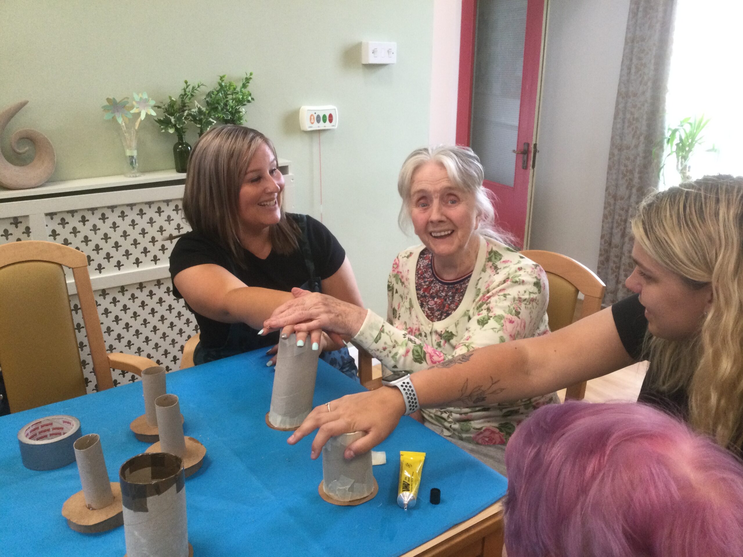 Three women seated side by side, their hands on top of cardboard tubes as they glue things in place