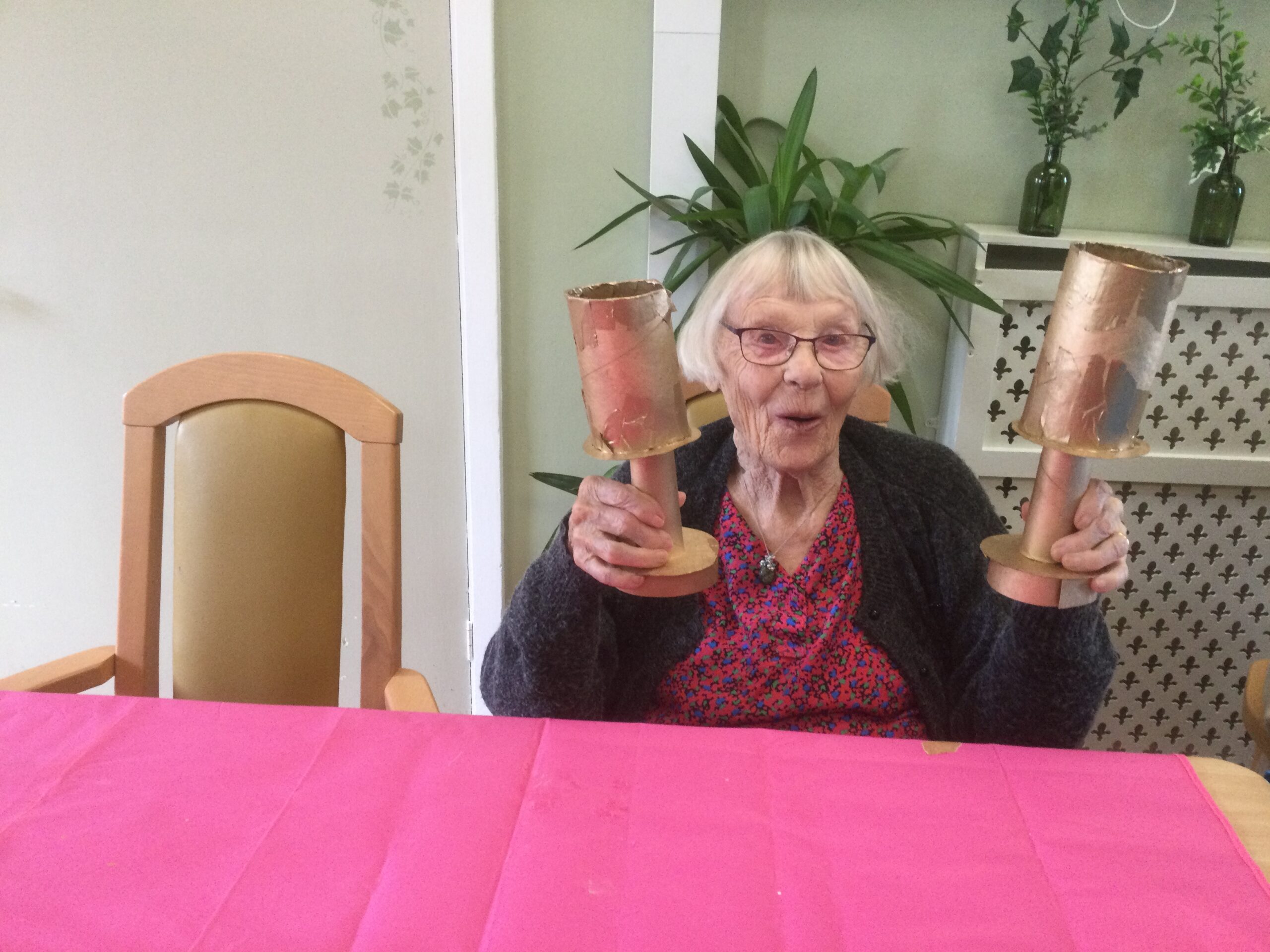 A woman seated at a table with a pink cover, holding up two cardboard trophies painted gold