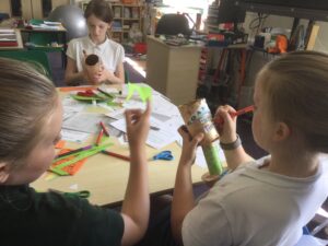 School children seated at a table, art materials in front of them, applying decorations to the cardboard trophies