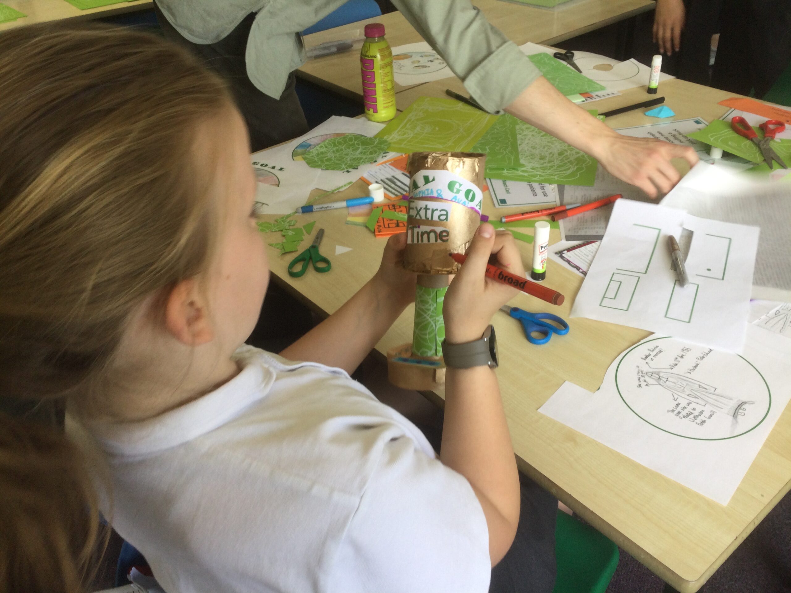 A table top covered with paper and art materials. A girl sits, writing something on the side of a cardboard trophy