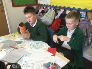 Two school boys in green tops seated side by side decorating a trophy with coloured papers and pens