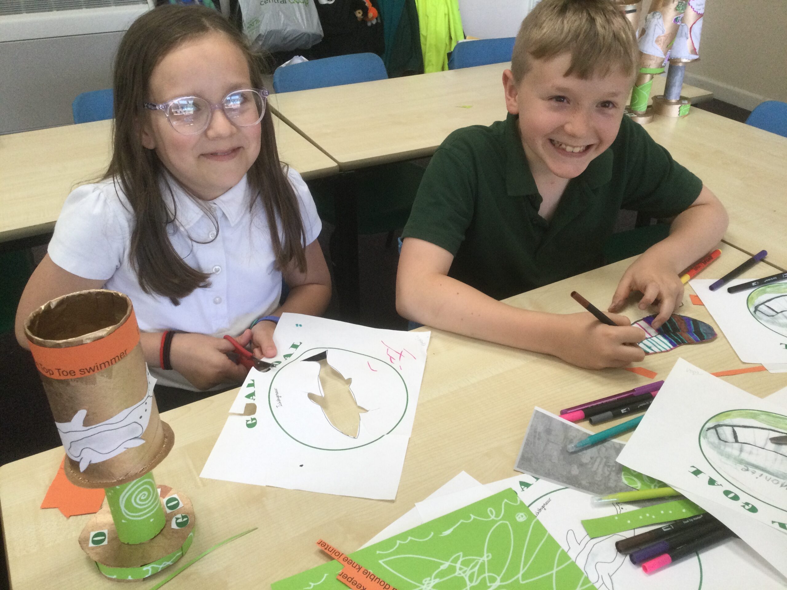 Two school children grin towards the camera as they cut out and colour in their art work