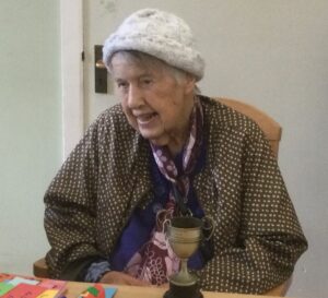 A woman wearing a woollen hat and brown spotted raincoat sits at a table, on which is a small silver trophy