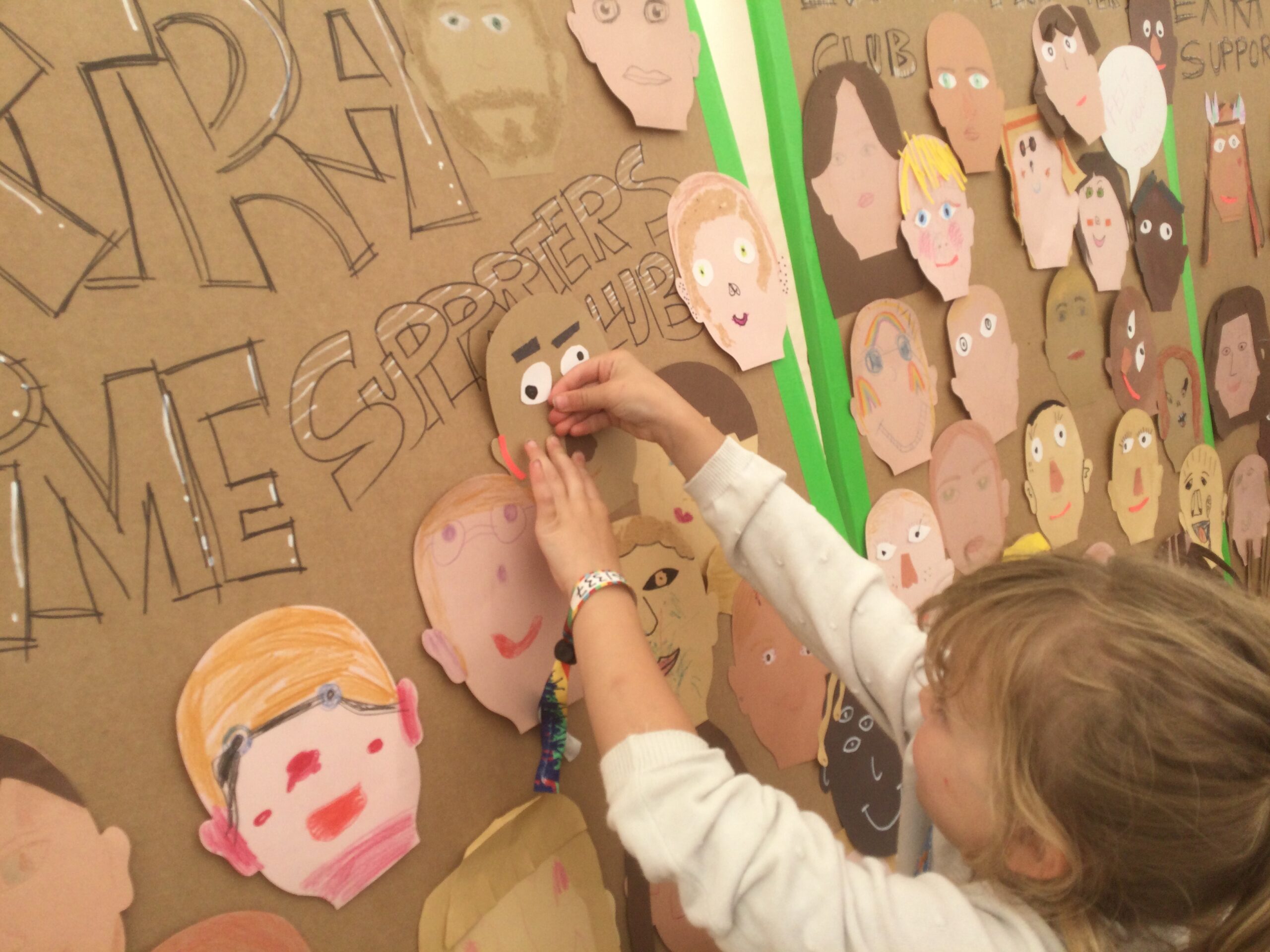 A child sticks a paper self-portrait to a cardboard panel on which there are numerous other faces