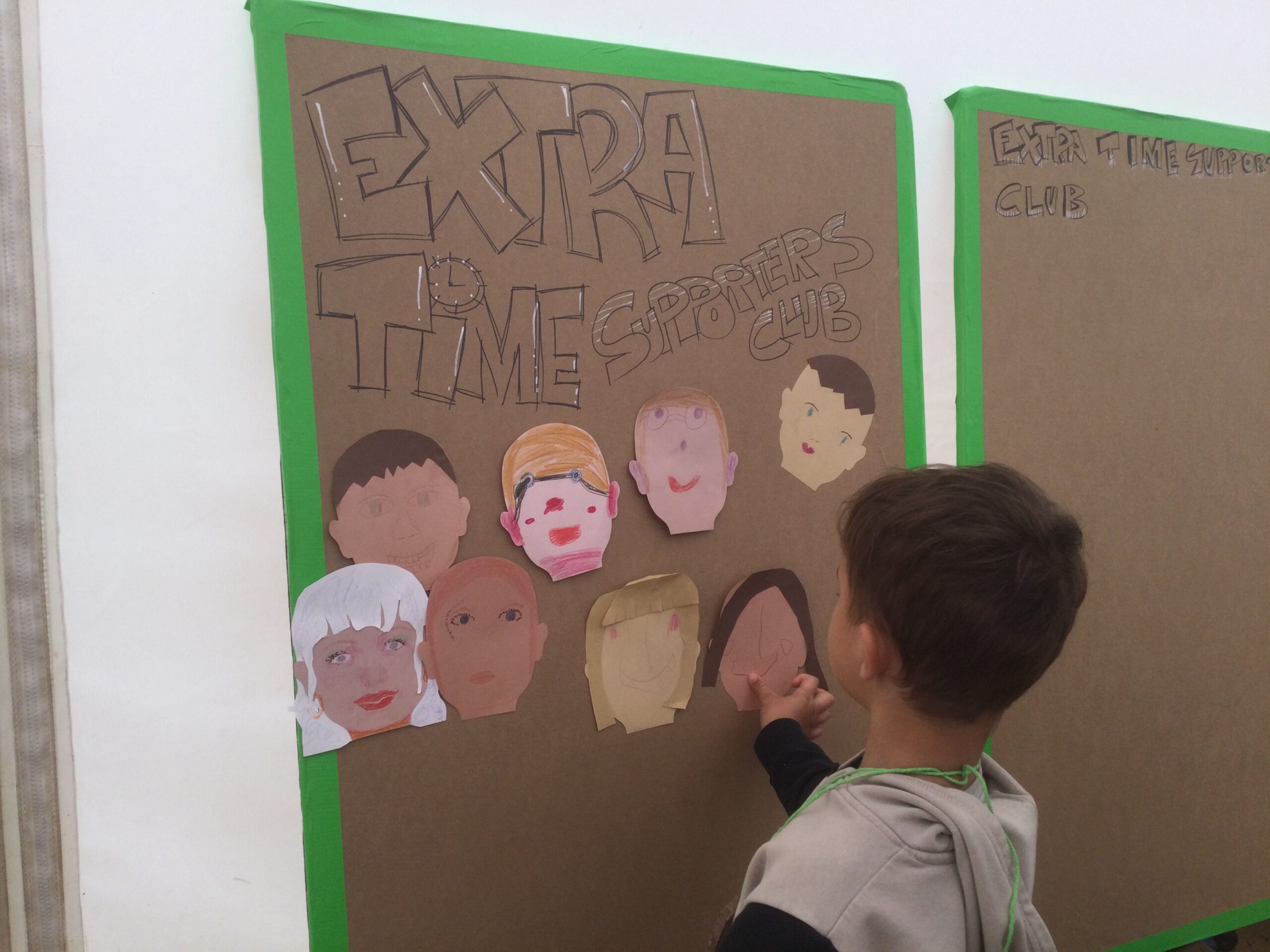 A cardboard panel with words Extra Time Supporters Club and rows of paper self-portraits with a young boy sticking up a face