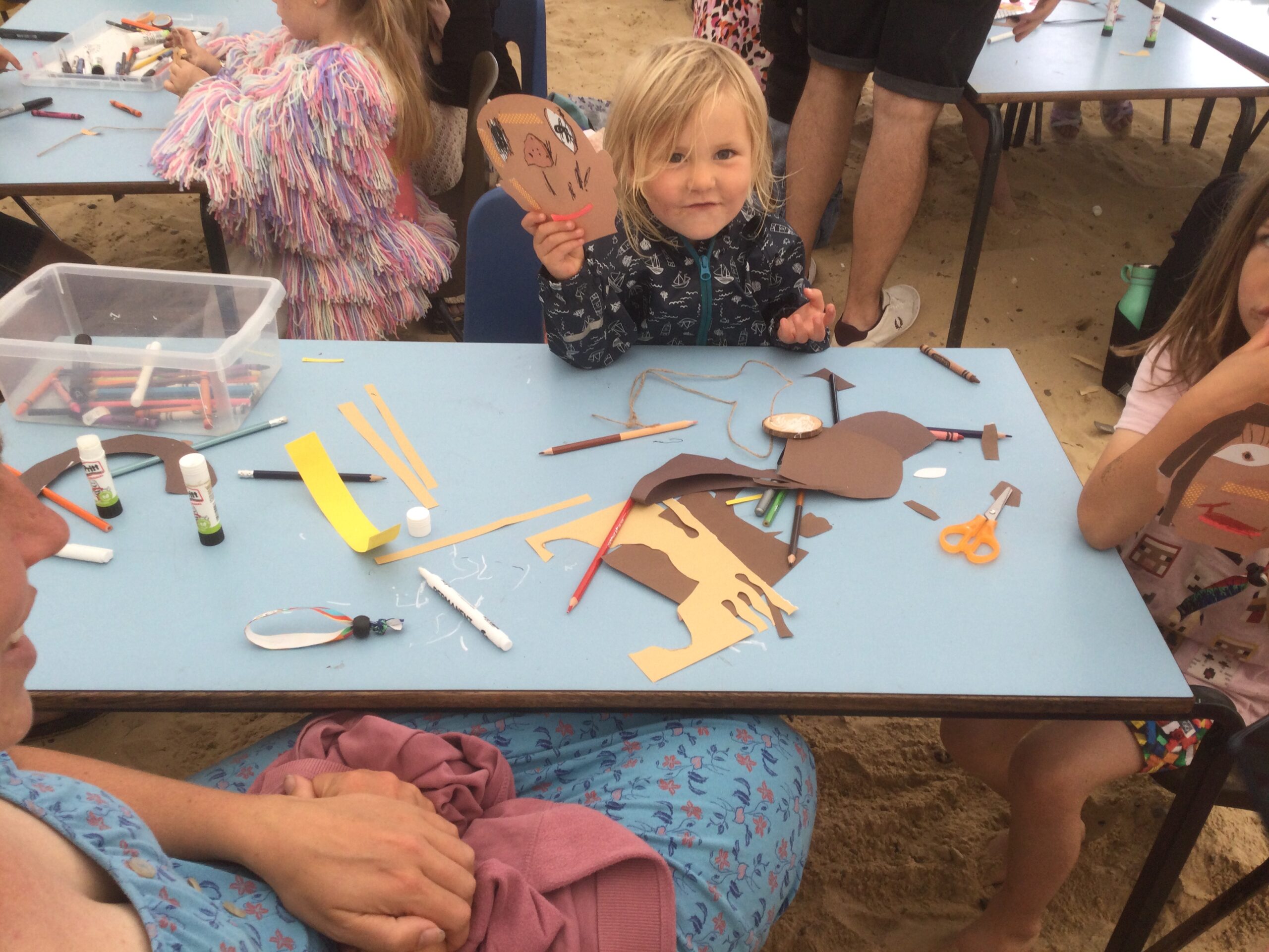 A child peeps out from behind a paper face whilst all around are art materials and coloured papers