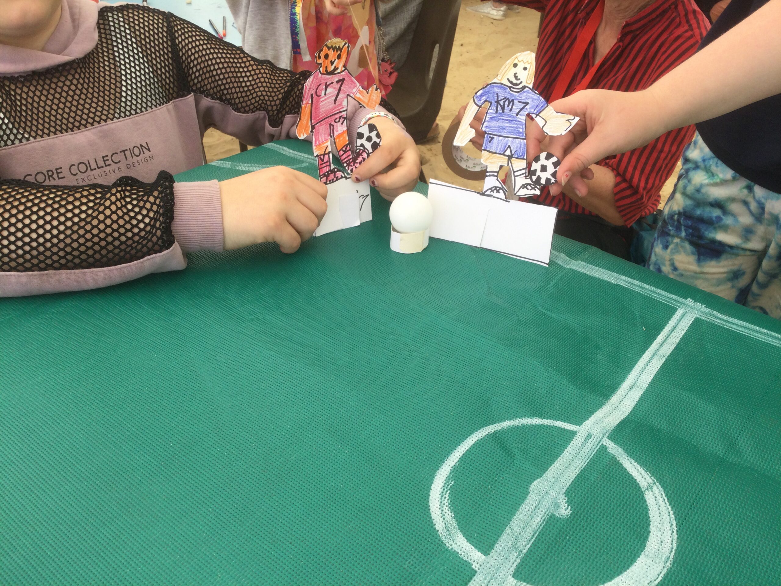 Children hold cut-out cardboard football figures on a green football pitch