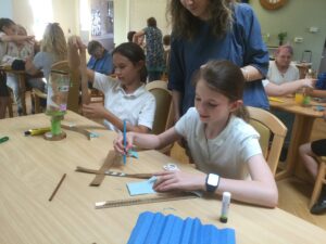 A busy room with adults and children all busy making things at their tables. In the foreground, two girls are cutting out strips of gold paper and gluing small blue hearts to them