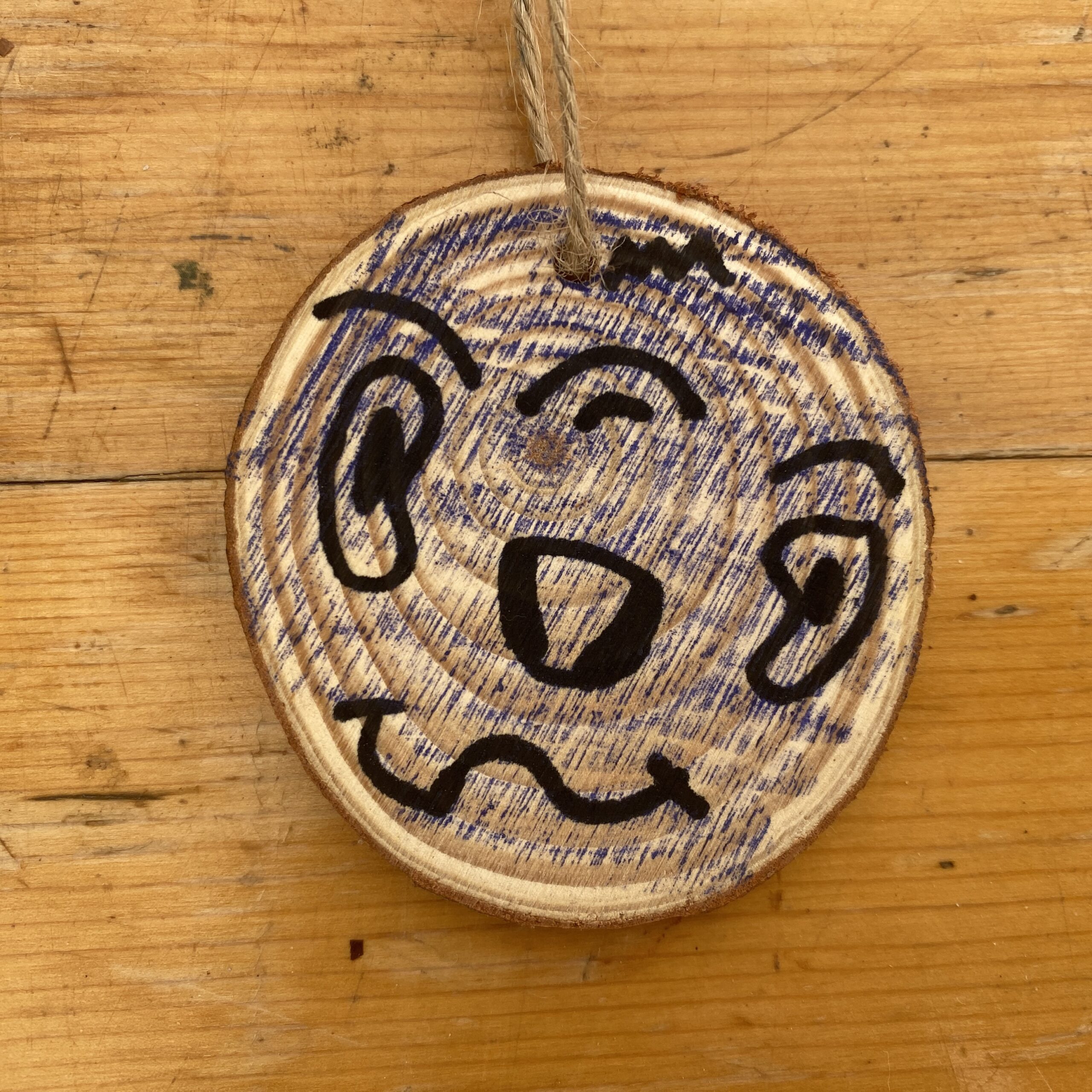 A round wooden disc with twine tied through the top and a face drawn in black felt tip pen with blue crayon background