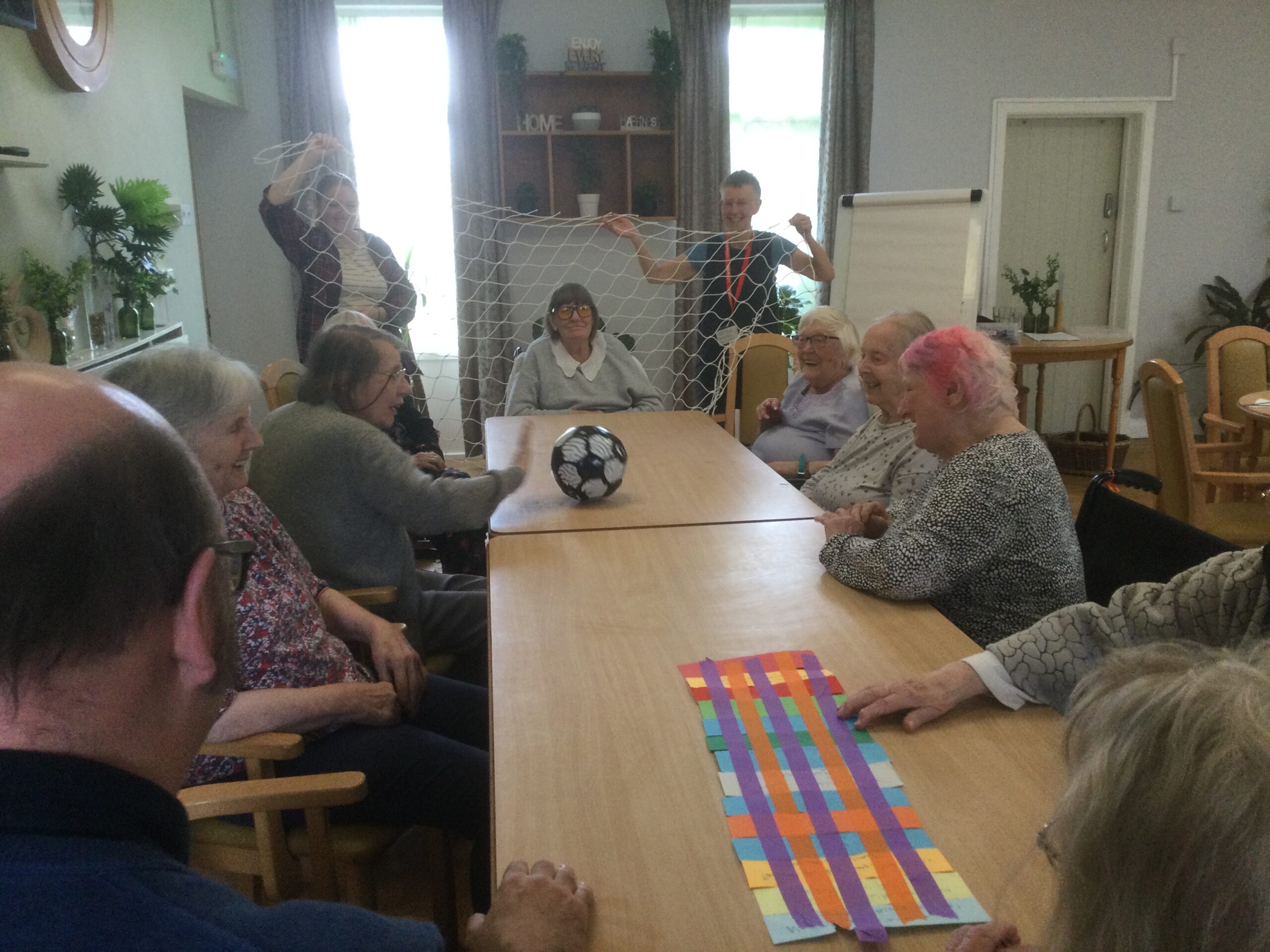 People sit either side of a long table, passing a black and white football back and forth. Behind the person seated at the end stand two people holding up a net like a football net. A multicolour piece of paper lies on the table nearest the camera