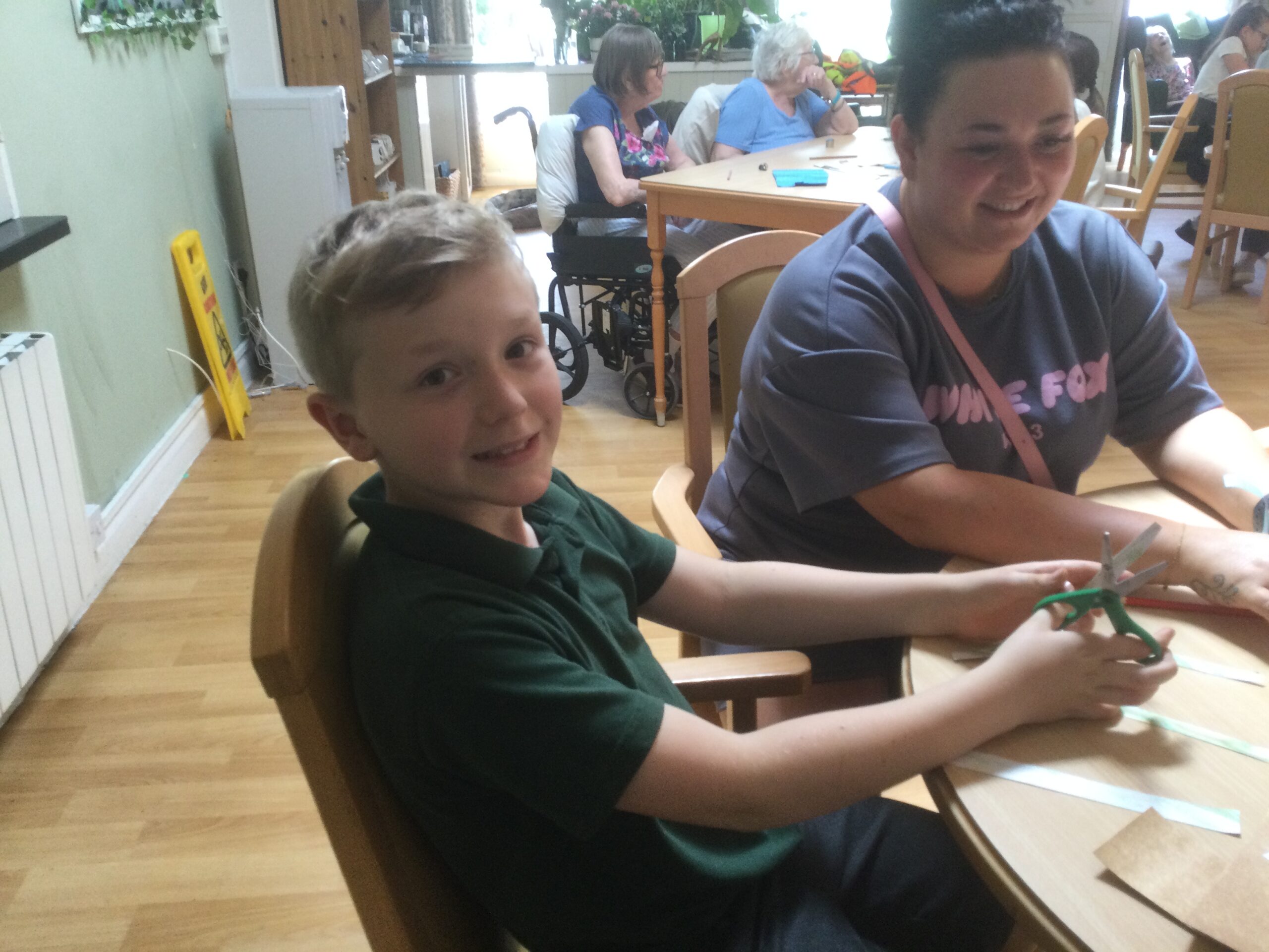 Foreground sits a young boy in a green school top, holding a pair of green handled scissors whilst beside him an adult in a blue top with pink writing on it smiles. There are people seated in the background