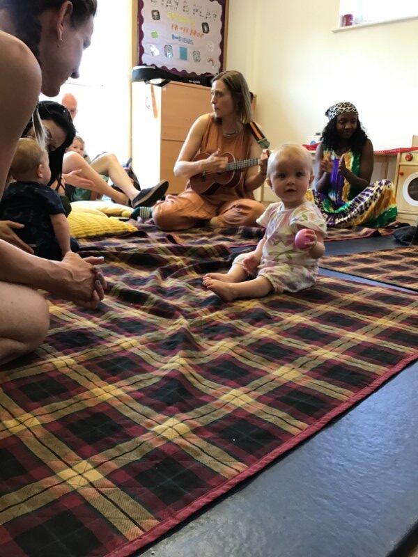 Charlotte Jolly and Kayleen Shani working with a group of parents and under fives. Everyone is sitting on rugs on the floor. In the foreground a child is smiling happily.