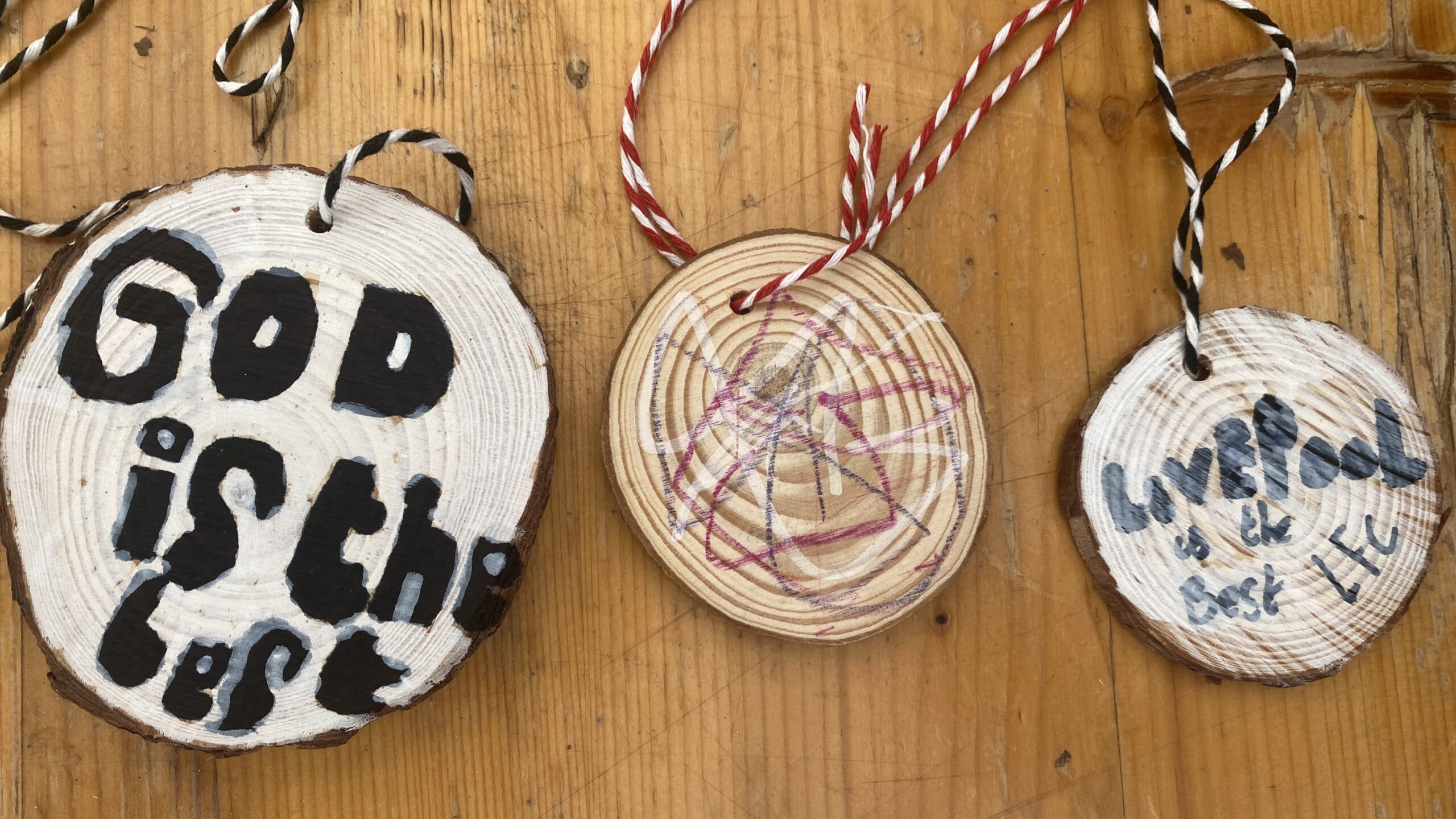 Three decorated wooden discs, each with twine through a hole in the top. One reads Good is the best and one reads Liverpool is the best