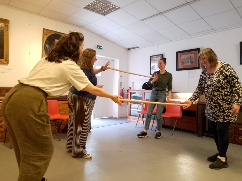 This photograph shows people in a workshop working in pairs to explore movement. Each pair has a pole which is held between them by each person pressing the palm of one hand against either end of the pole. You can see that the pairs are trying different movements while keeping the pole from falling by maintaining an even pressure on each end of the pole. The participants are concentrating and laughing a lot too.