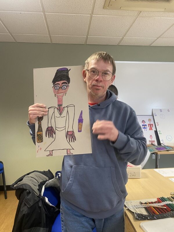 A photo of the artist holding his self-portrait in the style of a Roald Dahl witch