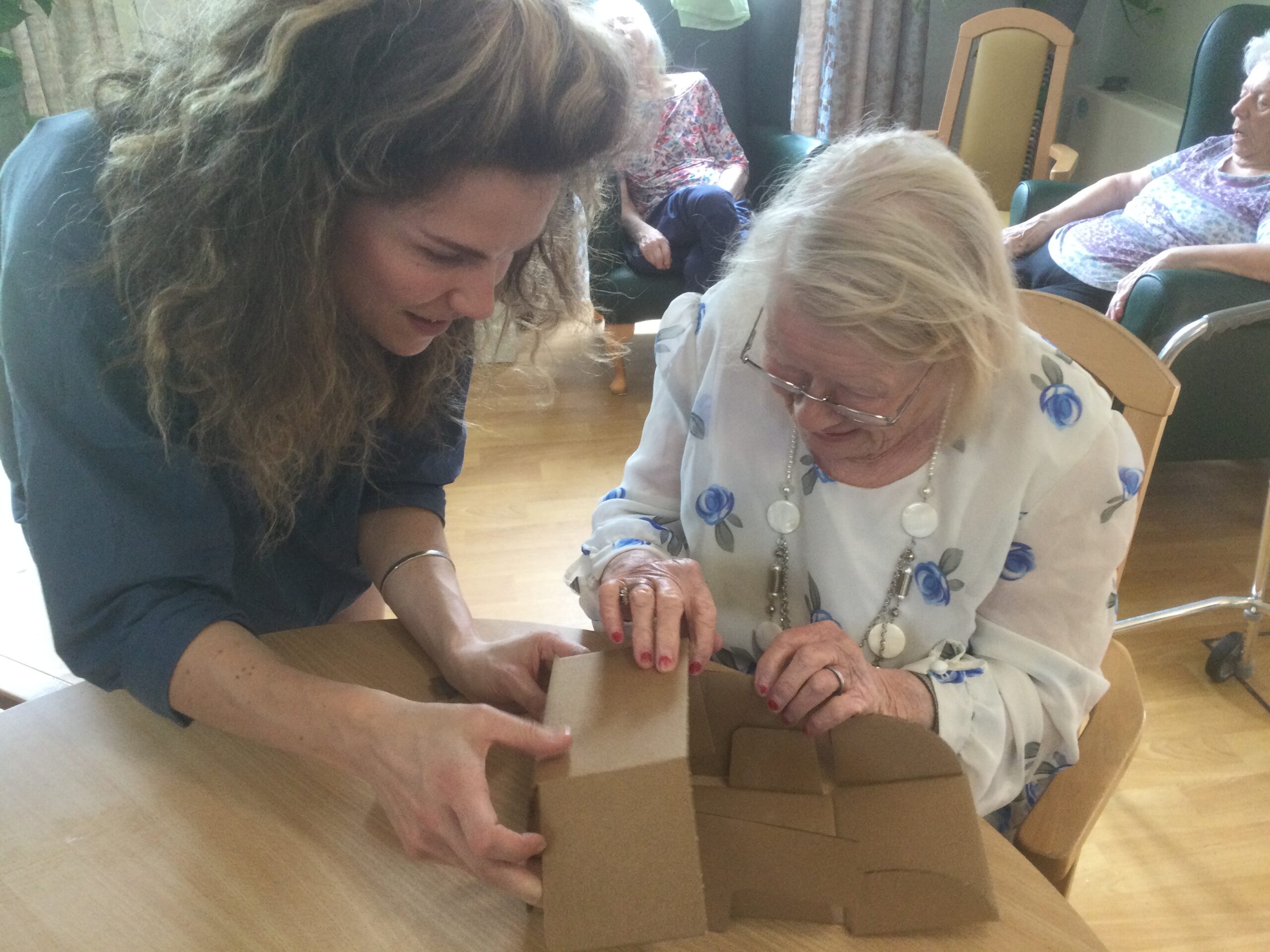 A woman in a floral blouse and with painted finger nails folds the side of a box whilst another person holds it all in place