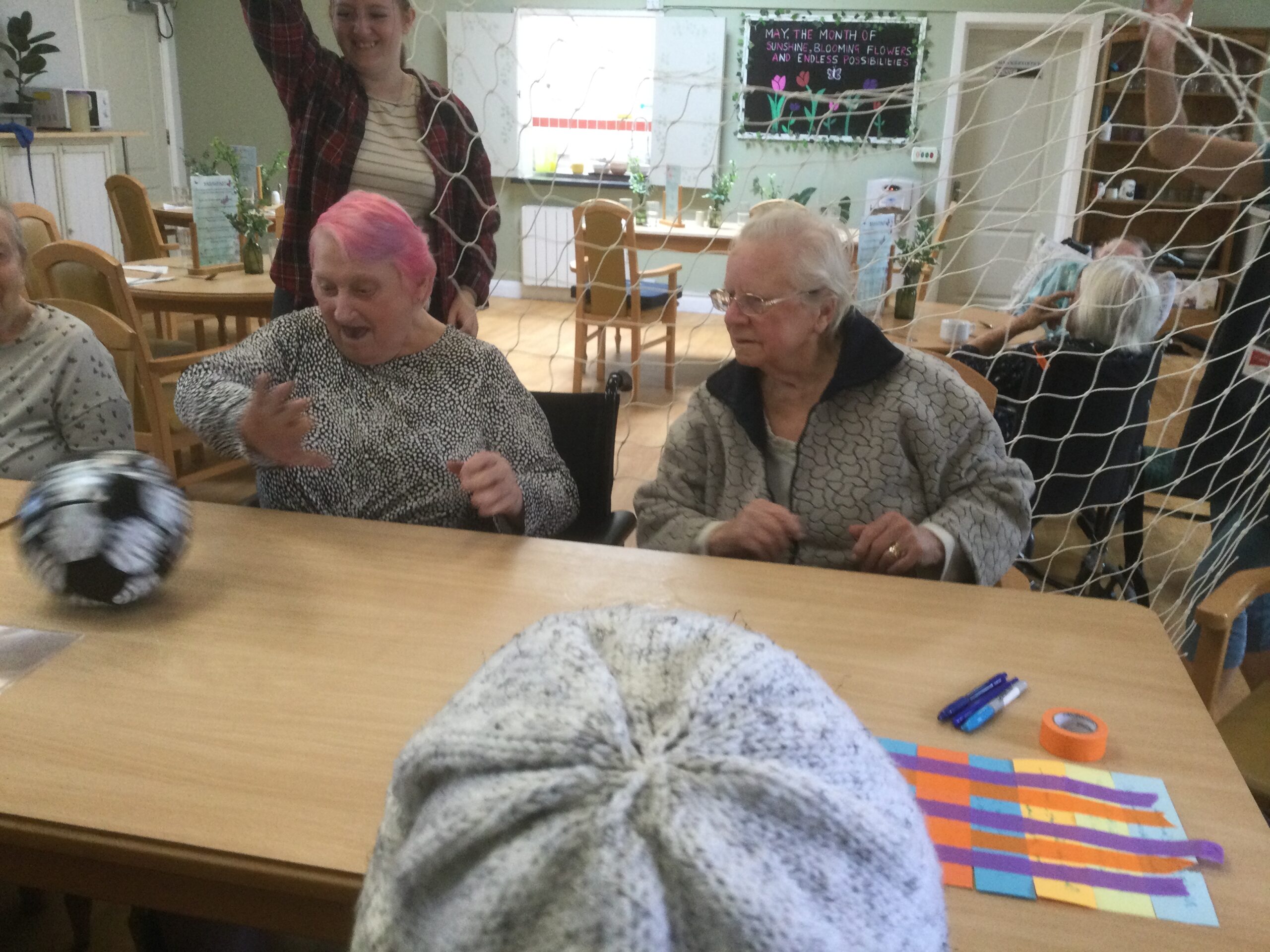 Two women flicking a black and white football across a table, whilst someone holds a goal net behind them.