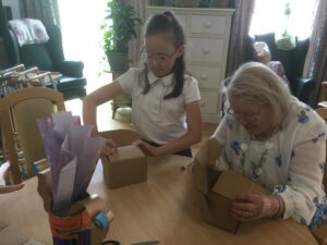 A school girl stands beside a seated woman, as both fold a flat piece of cardboard into a cube