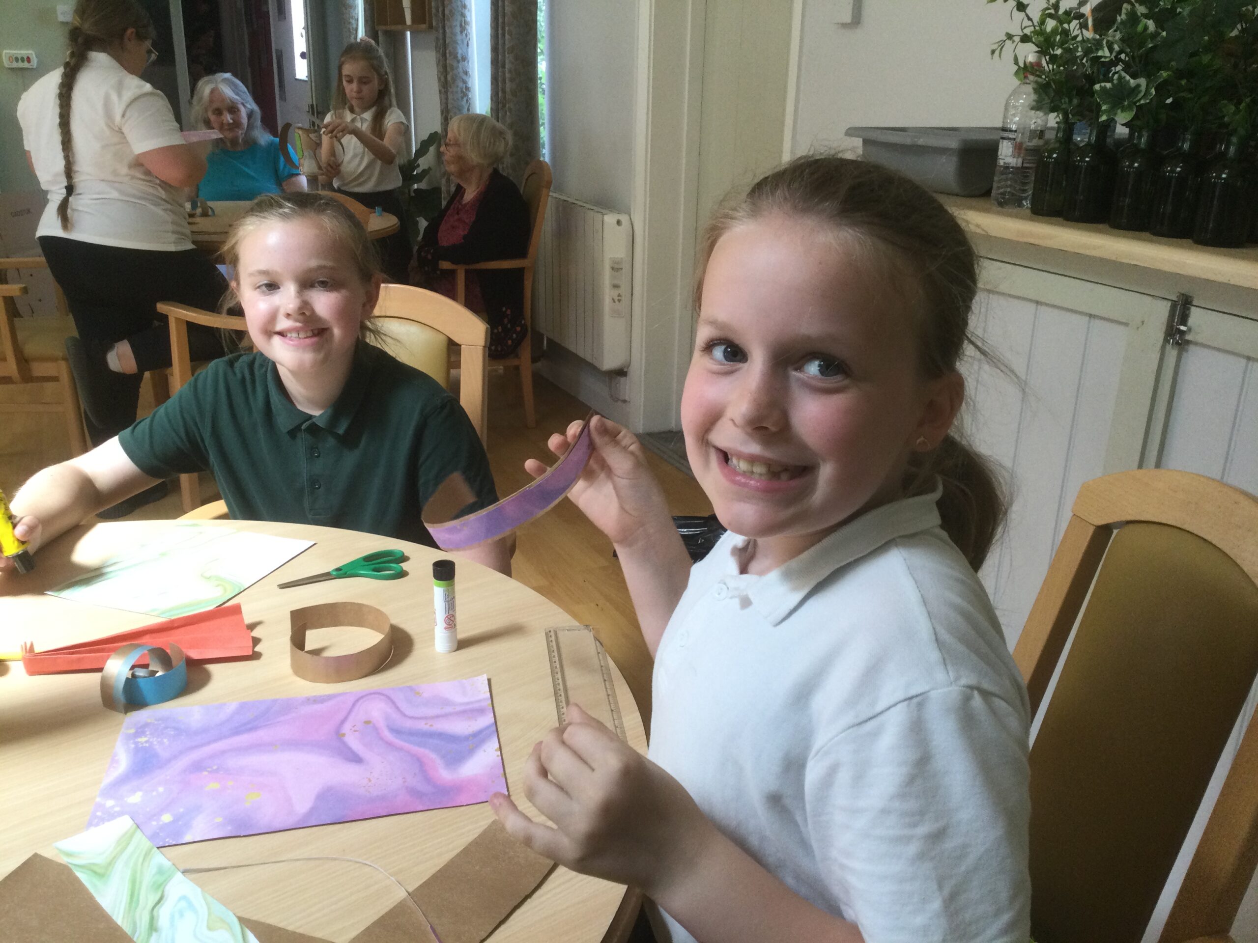 Two school girls beam at the camera, seated at a table covered in craft materials