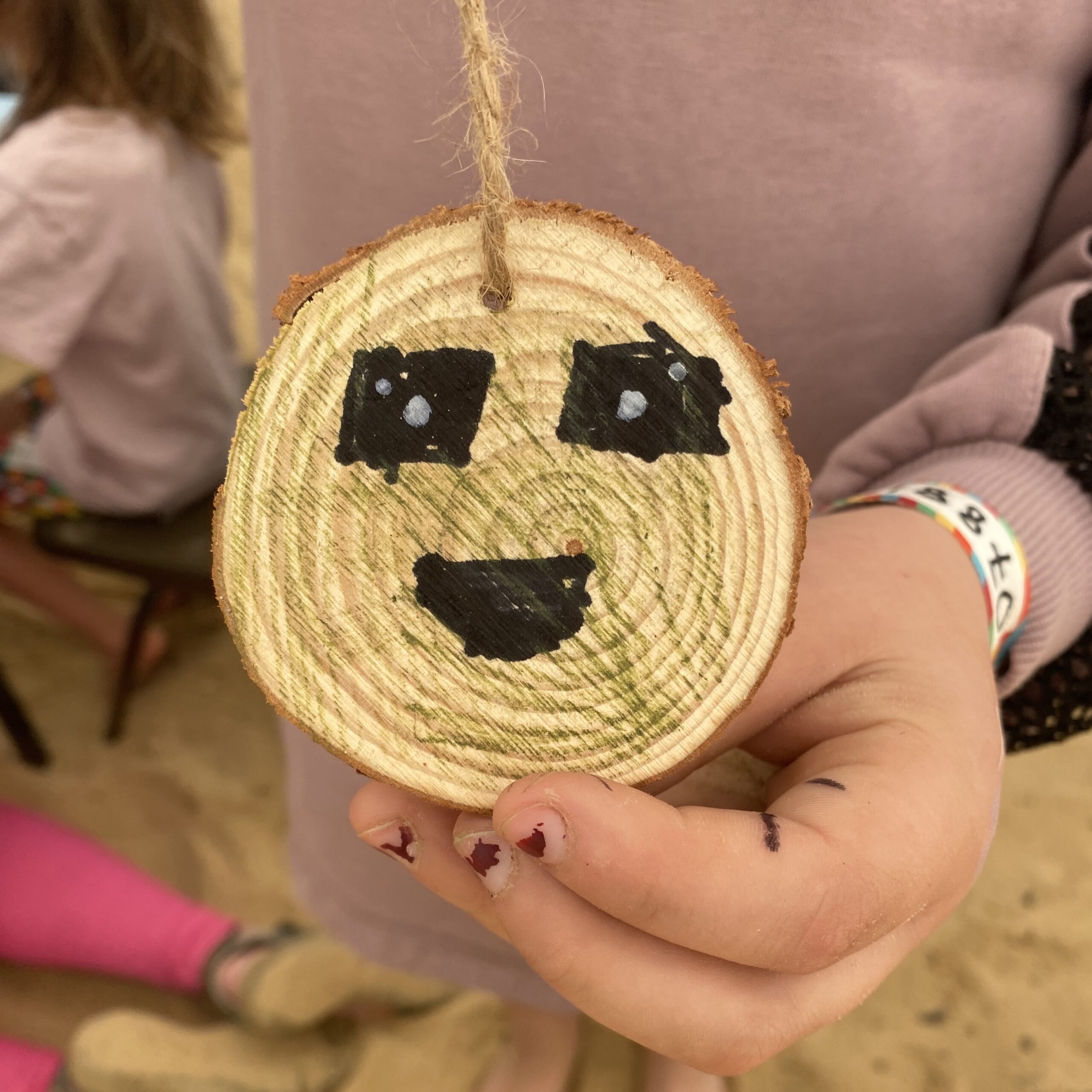 A child with painted finger nails holds up a wooden disc with large black eyes and mouth drawn on it