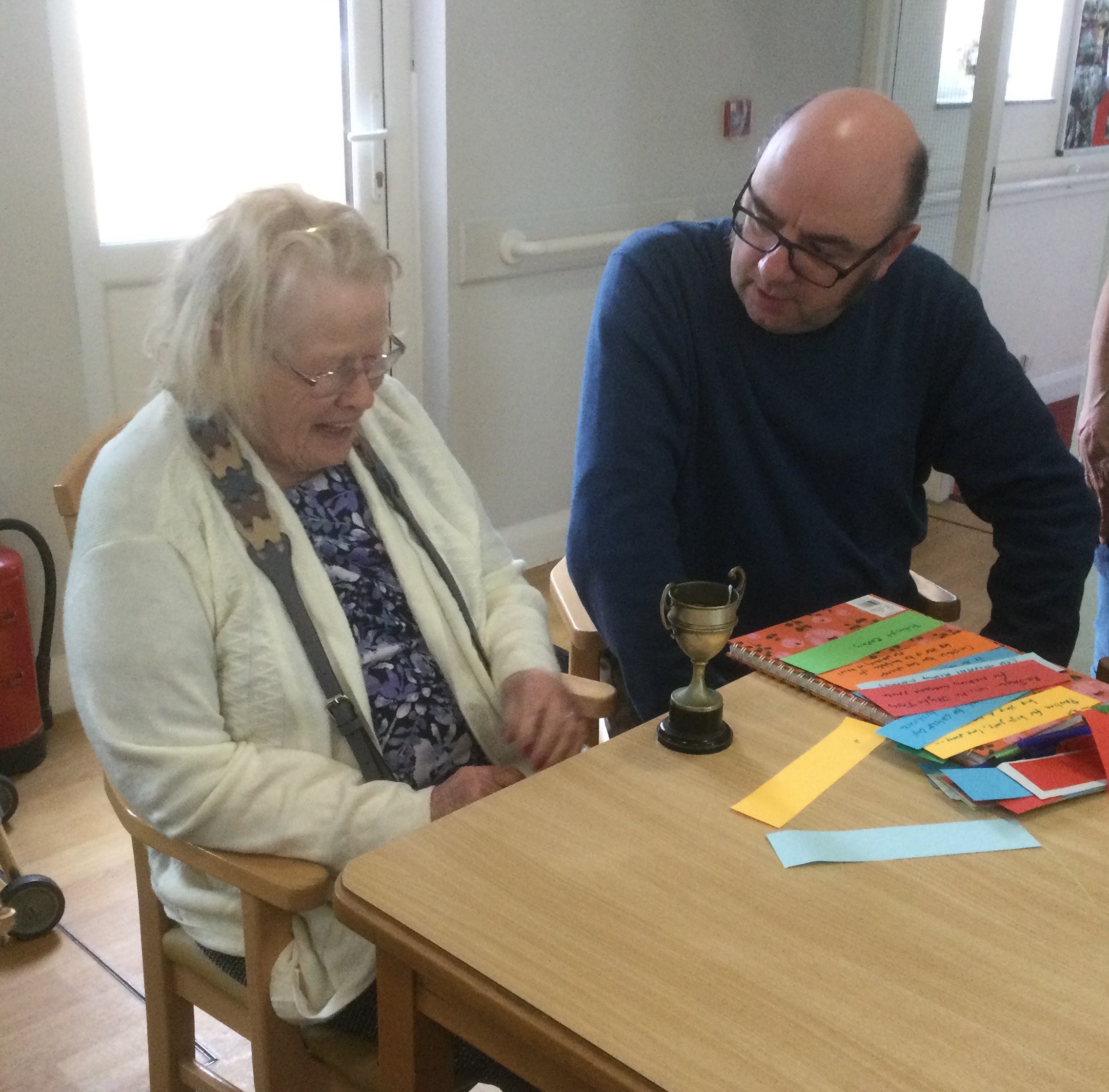 A woman sits looking at a small trophy on the table in front of her whilst a man, with spectacles and dressed in a dark jumper, engages her in conversation. In front of him are numerous strips of different coloured paper