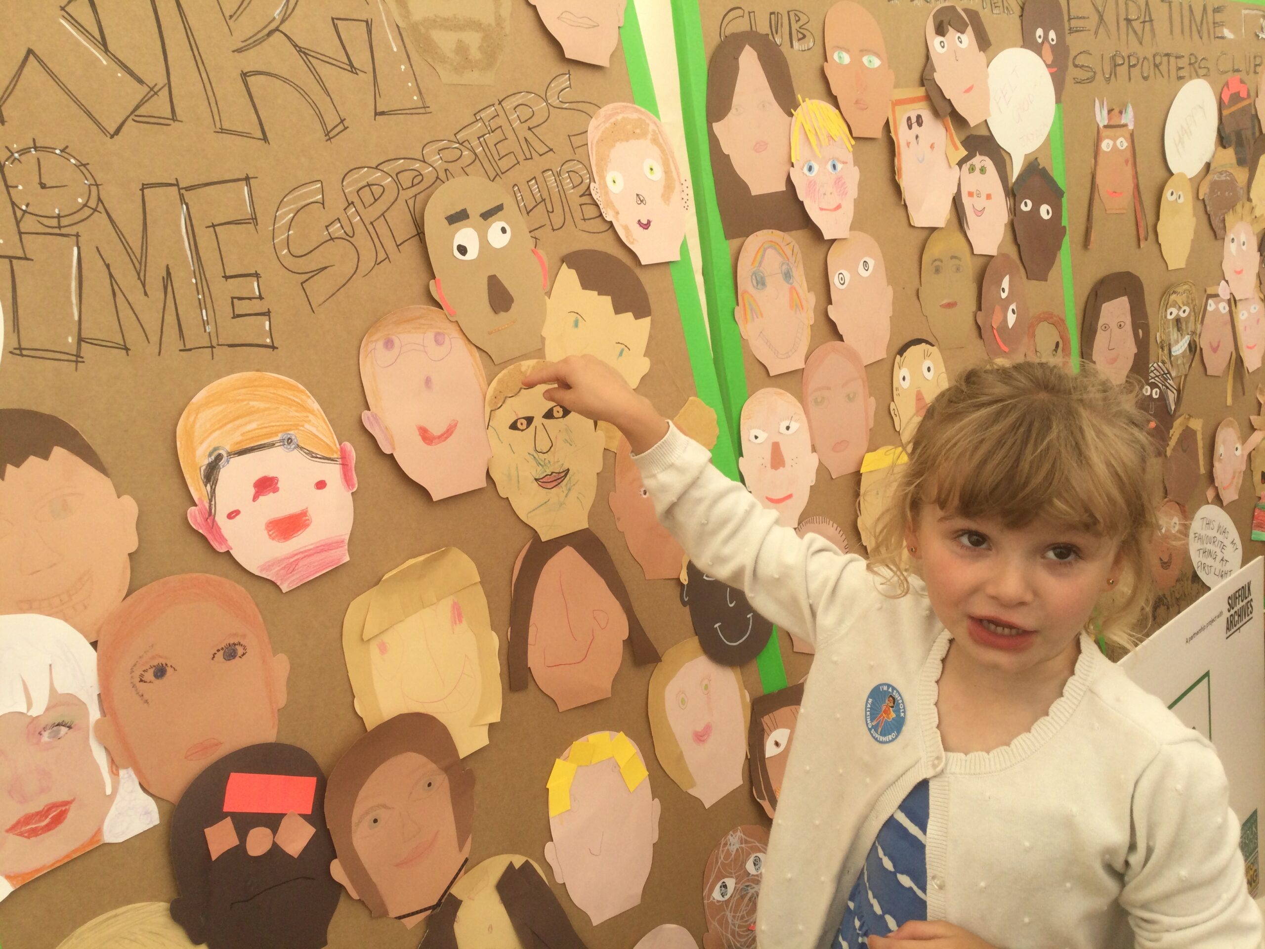 A child points to a paper self portrait stuck to a large cardboard panel, on which are many other faces