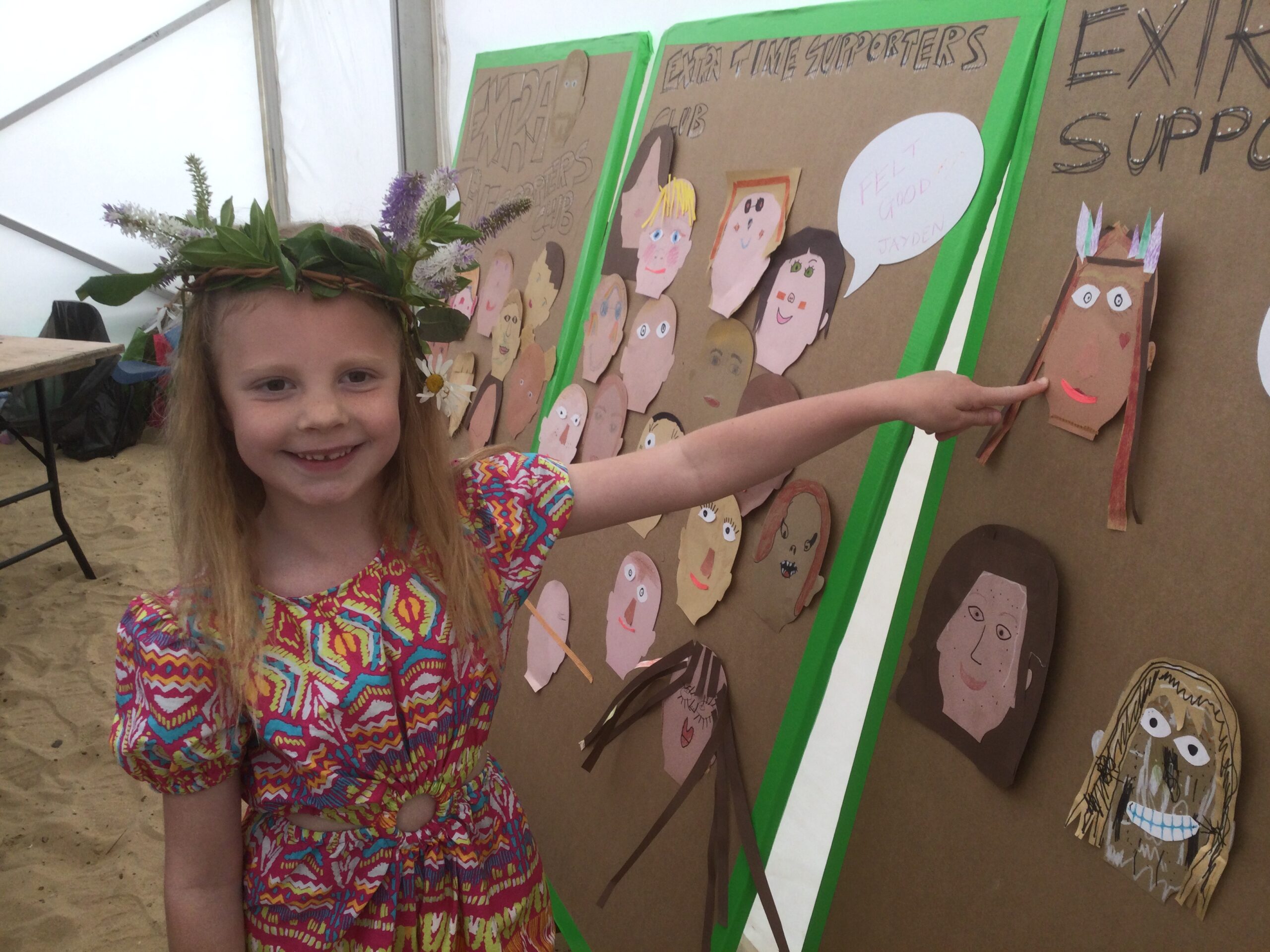 A child with a floral headdress points to a self-portrait of herself stuck on a large cardboard panel