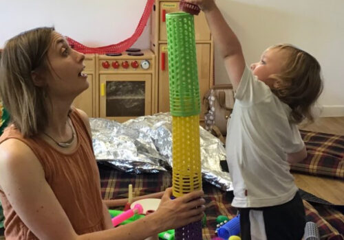 Charlotte Jolly and a child are sitting on rugs on the floor. They are building a tower from coloured,stackable, plastic cylinders.