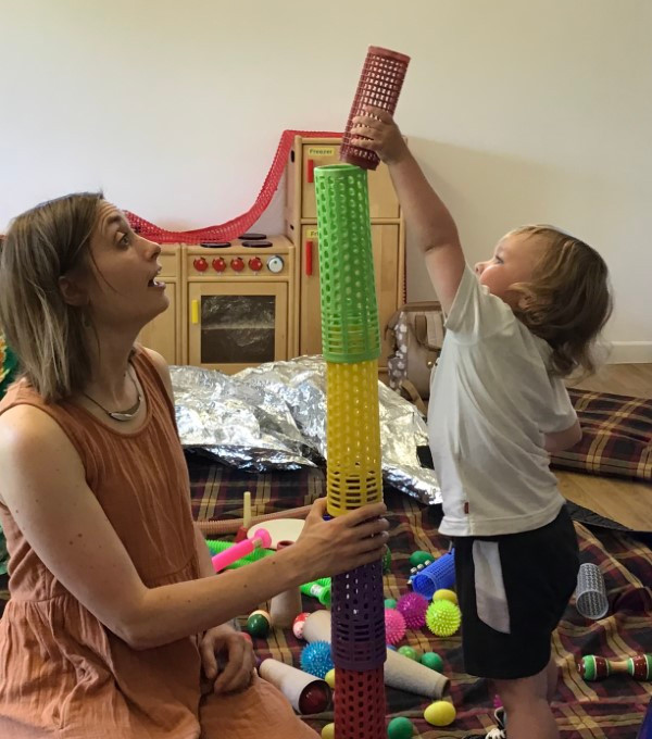 Charlotte Jolly and a child are sitting on rugs on the floor. They are building a tower from coloured,stackable, plastic cylinders.